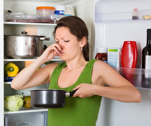 A woman holding her nose at spoiled food in the pot in front of the refrigerator