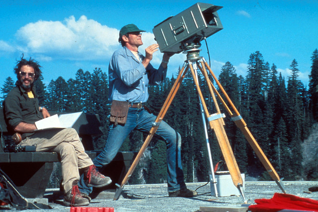 Image shows two geologists manning a geodimeter station. There is a line of conifers in the background. The geologist on the right, a dark-haired man with a beard, wearing a dark gray work shirt and khaki trousers with hiking boots, is sitting on a bench with his legs crossed, holding a folder with a page blowing over his hand. His hair is being tossed by the breeze. He is looking at the camera and smiling, looking very fly with his aviator sunglasses. The geologist next to him is wearing a blue workshirt and blue jeans, with a leather pouch hanging from his waist. He is standing in a very dramatic pose behind a piece of equipment mounted on a tripod which basically looks like a metal box with a flap lifted from the front. The man has a clean-shaven, jutting chin and looks almost like a blue-collar superhero. The whole scene gives off an almost Zoolander vibe.