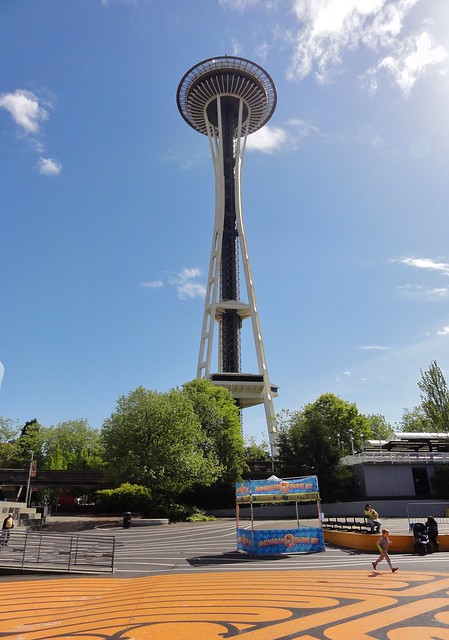 Image is looking up at the Space Needle, with the ribs of the underside of its disc visible. In the foreground, a portion of a labyrinth made of orange lines painted on asphalt is visible. There is a person in a gray sweatsuit energetically walking the labyrinth.