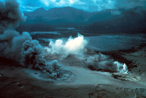 Image is looking across a plain of volcanic deposits towards mountains in the background. Everything is dark and gloomy except for the brilliant white steam plumes emerging from a crevasse in the foreground deposits. There is a crater with dark gray ash roiling from it in the center front: the ash plume is being blown toward the back left of the photo by the wind. Just beyond the plumes, scattered patches of dark water are visible in the destruction: the new Spirit Lake.