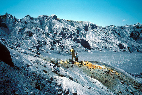 Image shows an amphitheater ringed by rocky gray ridges. There is a touch of orange in the rim of the far ridge. In the foreground, another, lower ridge is topped with a streak of yellow-orange. Two geologists stand on top of the long orange deposit, looking down into the smooth volcanic ash scattered with logs. One has his hands on his hips.