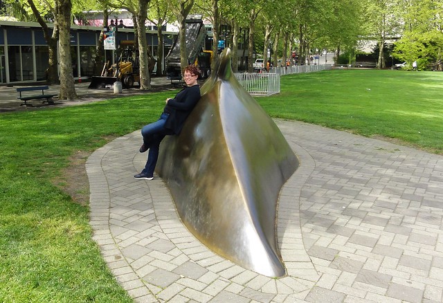 Image shows Aoife leaning her back against a bronze statue of a whale, which looks like it's swimming in the ground and only its top half has breached the surface. It's surrounded by brick paving. There is a green lawn around it.