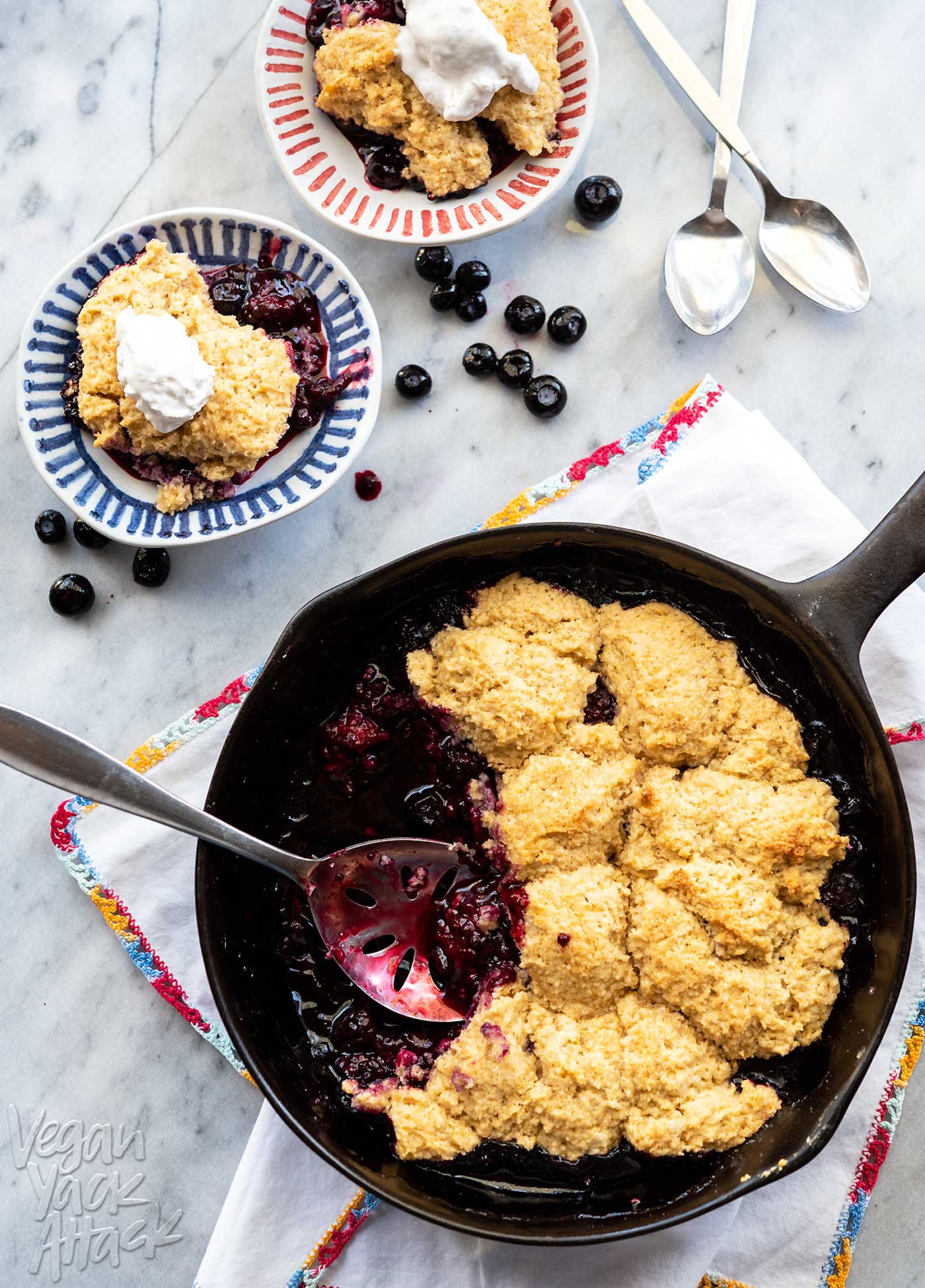 Cast iron skillet of blackberry cobbler with biscuit topping, with small plates of cobbler around it