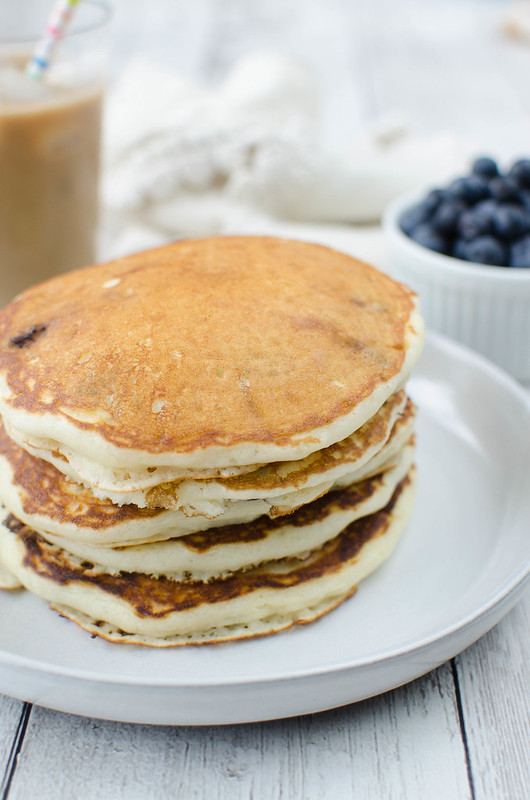 A stack of 5 blueberry pancakes on a white plate with iced coffee and a bowl of blueberries in the background