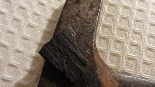 Image shows the head of a rock hammer. The bit of wood is resting against it; a shiny silver scratch shows by the fragment.