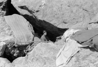 John Robert (Jack) Murrell, standing in a chasm between large boulders [bivouac site], with tent spread out on rocks above, Mount Tutoko, Darran Range, Southland Region