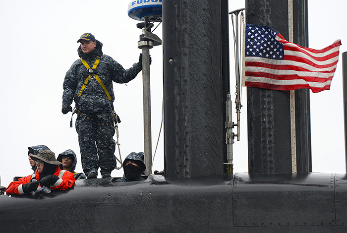 A man stands atop a submarine and holds onto a pole; the American flag flies to his right.