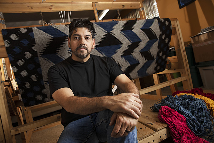 A man sits in front of a weaving loom that has a tapestry on it.