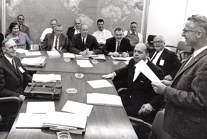 A group of people sit around a large desk; most of the image is black and white, but the only woman in the room is in color and wearing a purple outfit.