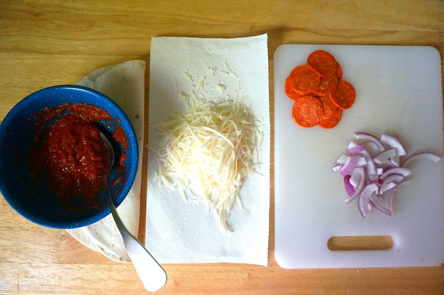 Pizza components laid out: a bowl of sauce sitting on a folded tortilla, a small pile of cheese on a paper towel, and small piles of pepperoni and red onion on a cutting board (protip: it's cute to fold the tortilla for a picture, but it makes it a little fragile, oops)