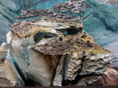 Image shows a plastic model of one of the glaciers that covered Washington State. The ice is varying shades of white and blue. It's cracked into deep crevasses. The top is covered with a mishmash of dirt and rock.