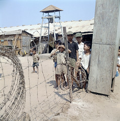 Fencing and barbed wire fencing with a Vietnamese refugee camp children and a large bell, Vietnam, 1967