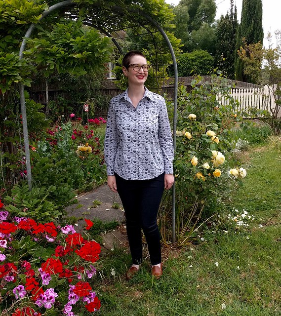 A woman stands in a garden archway. She wears a cutlery-print button-up shirt and dark skinny jeans.
