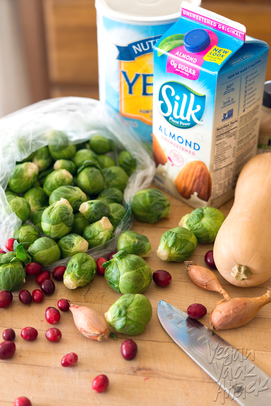 Fresh Brussels sprouts falling out of a bag, next to cranberries, shallots, butternut squash, nutritional yeast, a knife, and a carton of non-dairy milk, on a wood countertop.