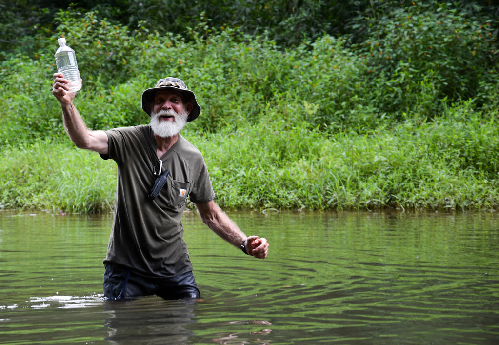 Jim Godwin standing waist deep in the water of Sugar Creek holding up a bottle of water collected as a sample