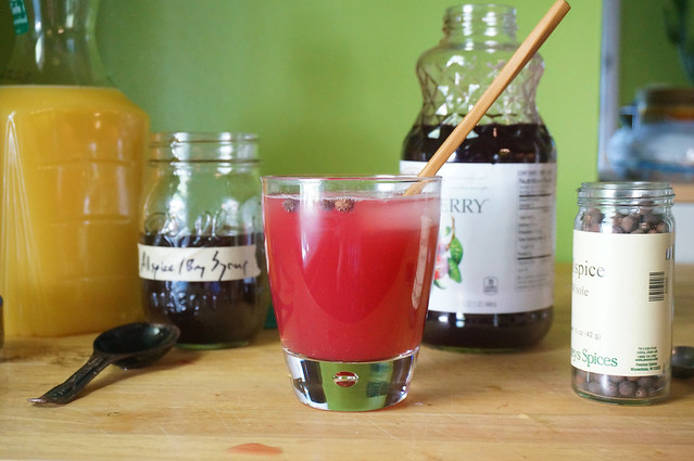 One serving of The Fizzing Cauldronsits on a wooden countertop, decorated with a few whole allspice berries and a bamboo stirring stick. Behind it, various bottles of ingredients: juices, syrups, spices.