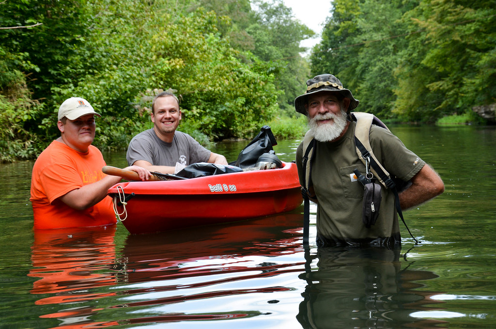Dustin Duffie Ben Wilson and Jim Godwin standing around an orange kayak in the waist deep waters of Sugar Creek surrounded by trees on either bank