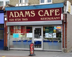 A small end-of-terrace shopfront with a blue, grey, burgundy, and cream sign above reading: “Adams Cafe / 020 8726 7865 / Restaurant”.  Below is a glazed frontage with multiple posters and notices in the windows, and an illuminated red sign reading “Open”.