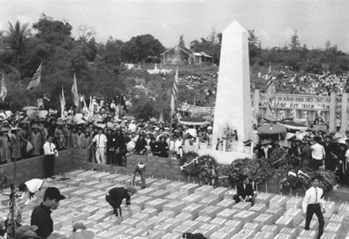 Populace watches as officials arrange coffins in front of memorial obelisk.