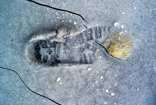 Image shows a hiking boot imprint in ash deposits. A small fumarole has spilled yellow-orange deposits over the tip. Diagonal cracks have formed in the deposits, cutting across the print.