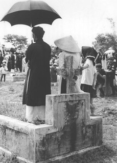 Elderly couple standing under umbrella in cemetery.