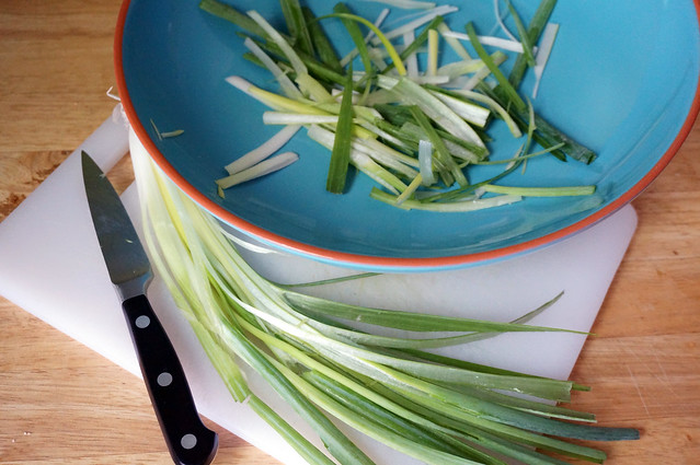 Two slivered scallions lie on a cutting board, curving around a bowl that already hold some smaller pieces. They look like a horse's mane of onion. A pungent, pungent horse's mane.