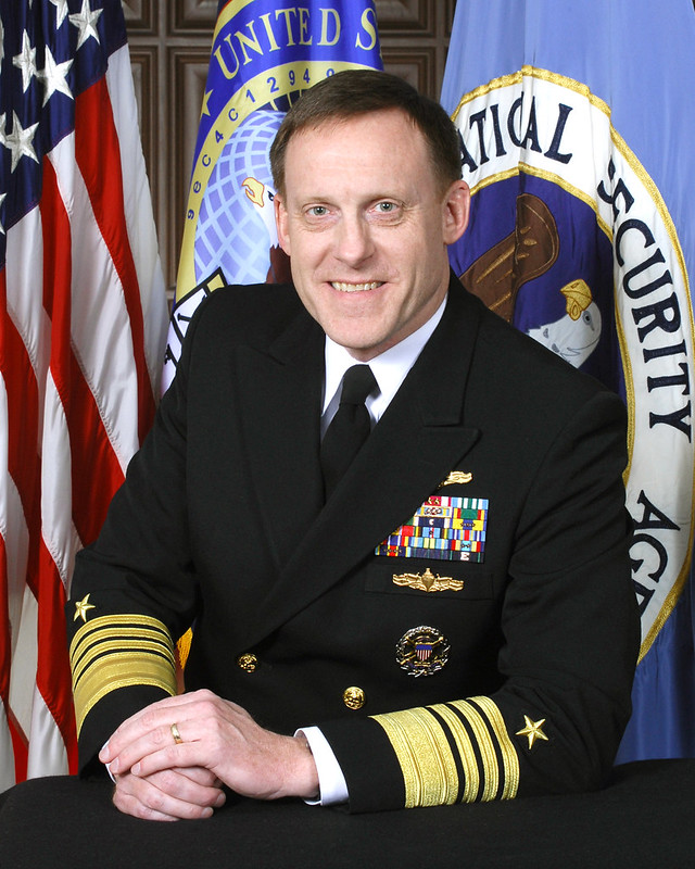 Navy Admiral Michael S. Rogers sits in front of three flags.