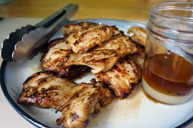 A plate heaped with slabs of richly browned chicken thighs. A pair tongs rest at the edge, the handle receding into the background, and a small glass jar filled with layers of pale green lime juice and tawny brown olive oil sits on the plate, chicken juices pooling around its base.